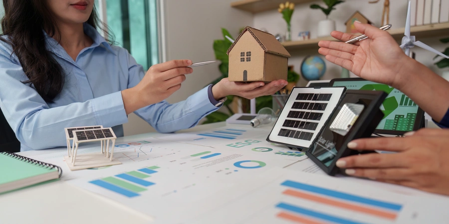 Consultant presenting a sustainable home plan with a cardboard house model and solar panel samples on a desk.